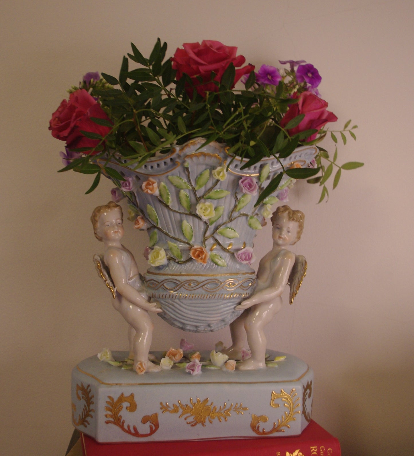 Capodimonte-style duck-egg blue vase with gilded cherubs and raised floral detail, styled on a vintage book with red roses