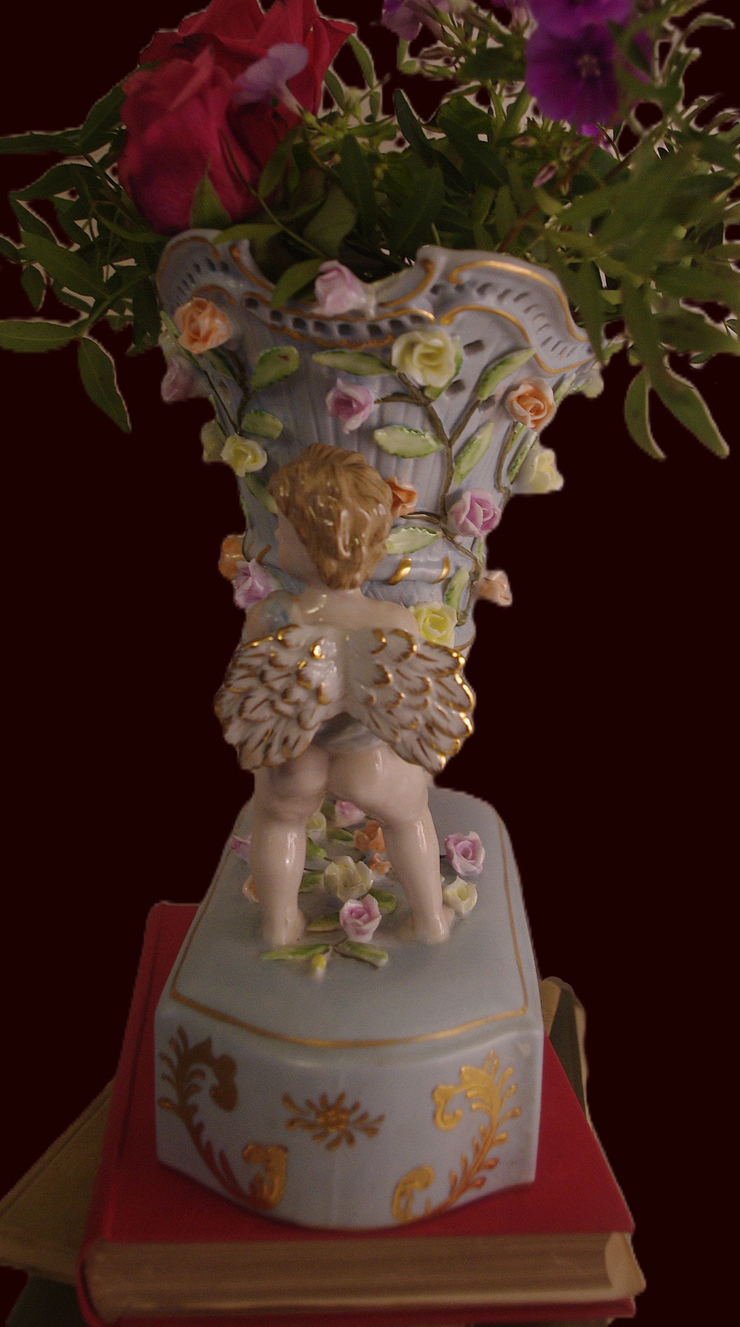 Capodimonte-style duck-egg blue vase with gilded cherubs and raised floral detail, styled on a vintage book with red roses and shown in side view