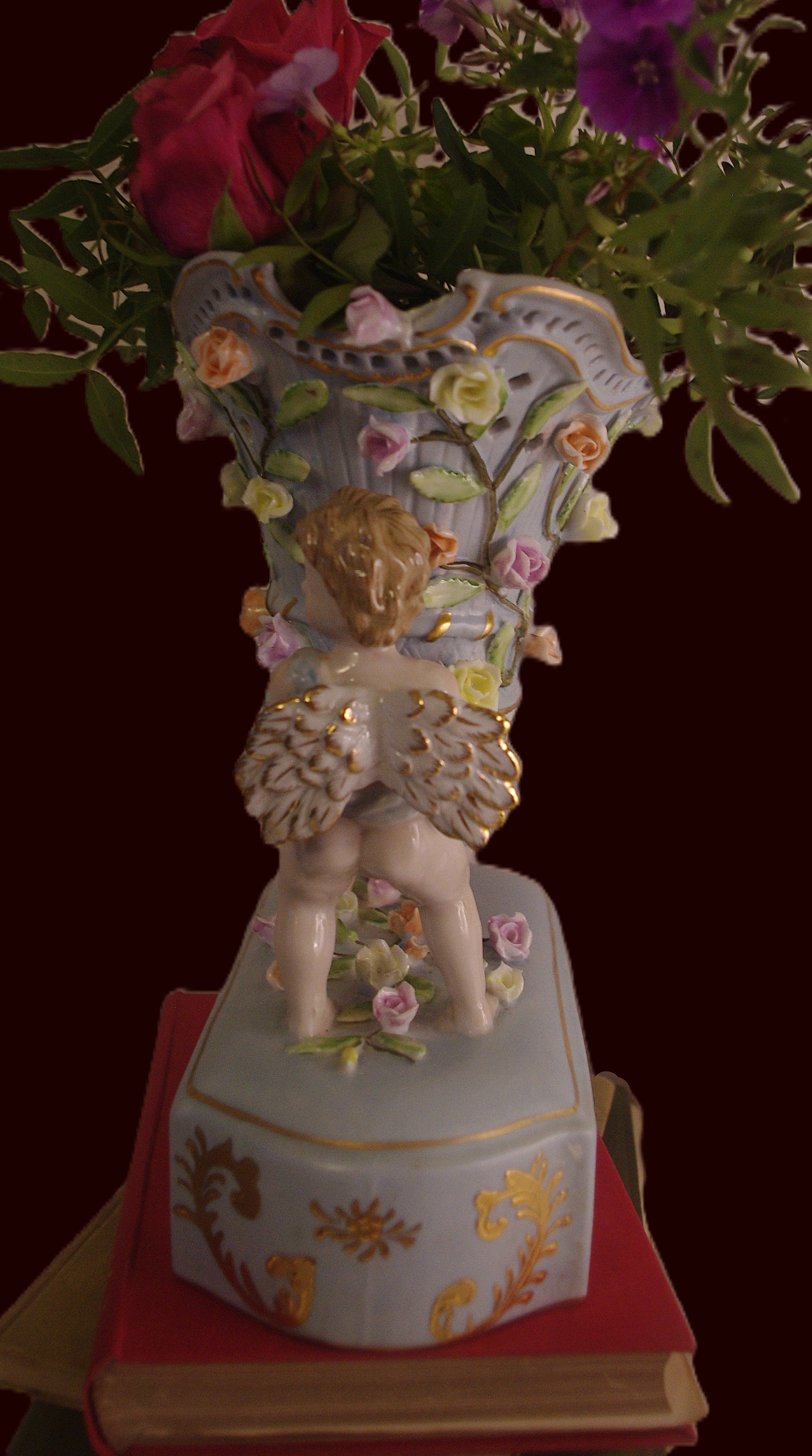 Capodimonte-style duck-egg blue vase with gilded cherubs and raised floral detail, styled on a vintage book with red roses and shown in side view