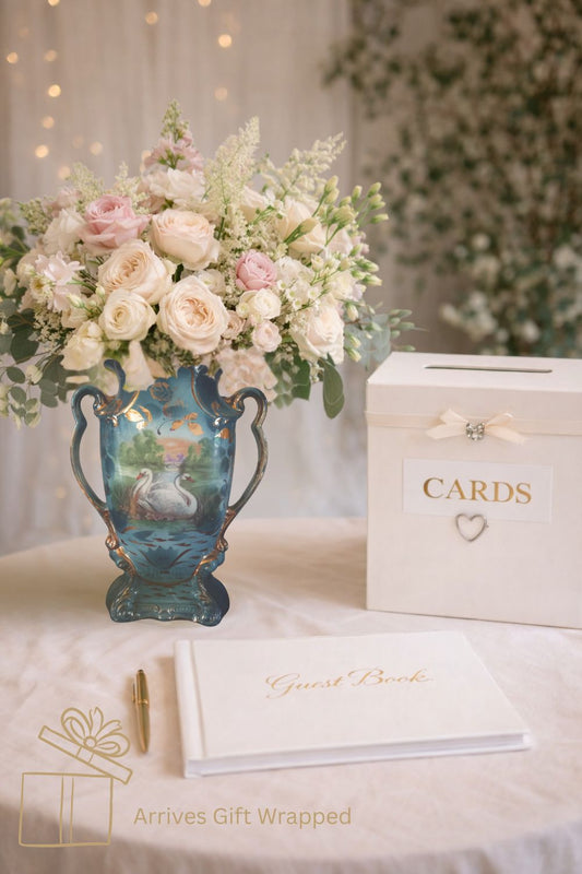 Romantic wedding guest book table featuring an antique English swan vase filled with blush and ivory roses beside a white wedding cards box and elegant guest book, styled for heirloom wedding decor