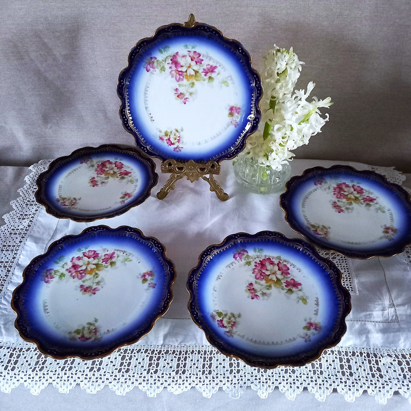 Five antique Limoges dessert plates displayed with one on a brass stand, Belle Époque cobalt blue rim porcelain with floral decoration and lace table styling.